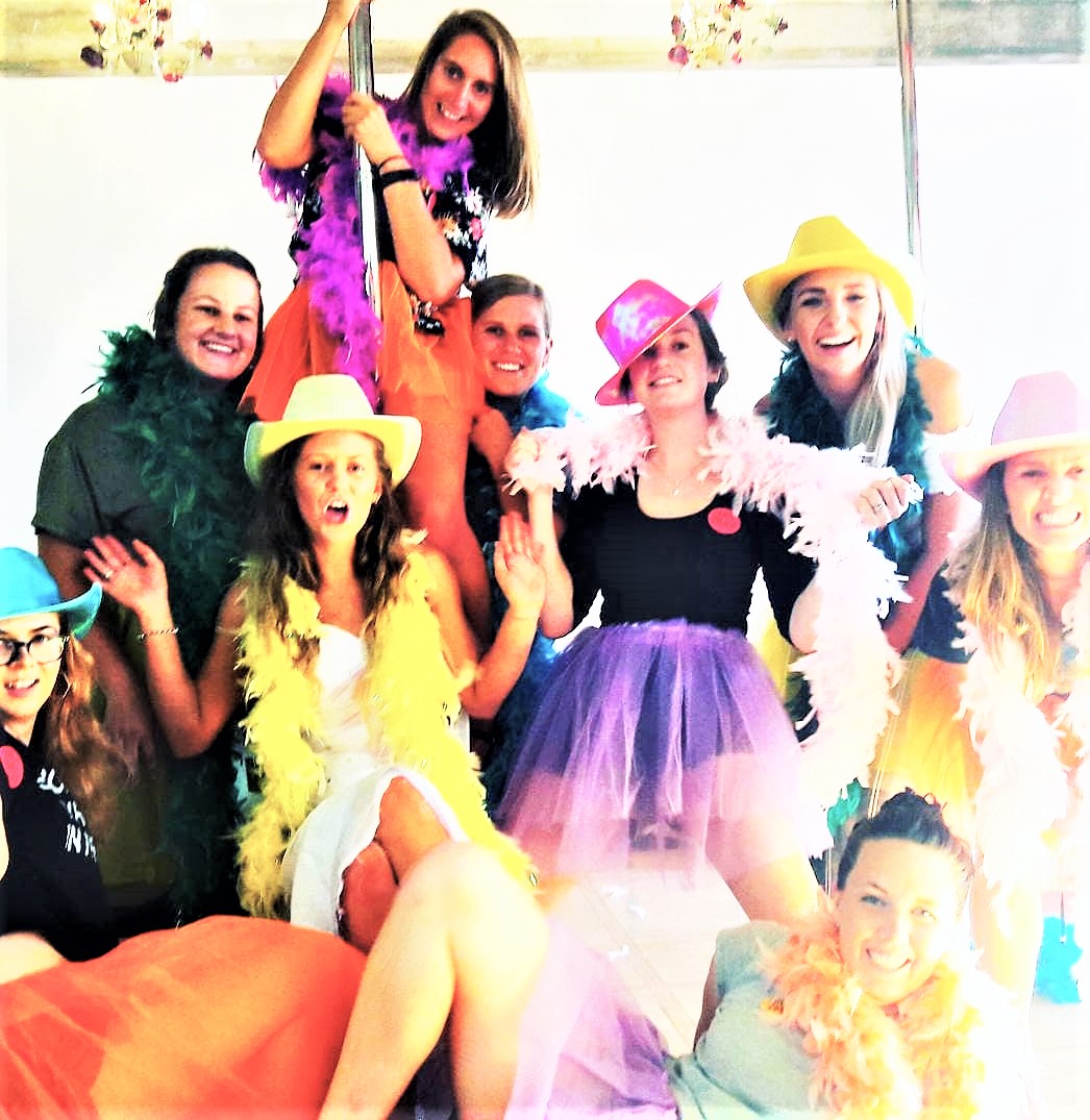 A group of smiling hen's night participants pose around a bride to be sitting in front of a pole dance pole. They are wearing colourful feather boas and hats.